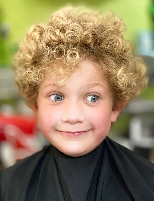 A smiling child with blond curls wearing a dark salon cape